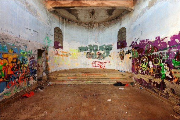 Fotosession in der Geisterstadt Es ist noch gar nicht lange her als ich bei www.fotocommunity.de einige Fotos einer Geisterstadt auf Teneriffa gefunden habe. Die Location schien wirklich cool zu sein und ich trage nun schon seit vielen Monaten den Wunsch mit mir herum, diese Geisterstadt mal genauer unter die Lupe zu nehmen. Aber was hat es mit dieser Geisterstadt auf sich? Als ich in den 60er Jahren das Licht der Welt erblickte wusste ich noch nicht wie froh ich sein durfte, dass meine Mutter während sie mit mir schwanger war keine Medikamente eingenommen hat. Bei tausenden anderer Menschen in meinem Alter sah das anders aus. Da gab es ein Medikament der Grünenthal GmbH, das besonders gut verträglich sein sollte und rezeptfrei in den Apotheken erhältlich war. Viele schwangere Frauen nahmen es gegen die morgendliche Übelkeit sowie Unruhe und Schlafstörungen ein und brachten einige Monate später schwer missgebildete Kinder zur Welt. Menschen bei denen die Hände ohne Arme direkt oben an den Schultern angewachsen waren. Viele leben bis heute mit diesen schweren Behinderungen und haben bedingt durch ihre Behinderung andere Krankheiten bekommen. Oft sind es schwere Rückenbeschwerden. Das Medikament hiess „Contergan“ und der Rechtsstreit der aus diesem ersten großen Medikamentenskandal der Menschheitsgeschichte hervorging, zog sich über viele Jahrzehnte hin. Eine Weile nach dem Skandal gab es den Arzt Dr. Jacob Sheskin der sich in Jerusalem um Menschen gekümmert hat die an der Lepra erkrankt waren. Heute kennen wir dieses Krankheit nicht mehr, aber als sie noch auf dieser Erde gewütet hat wurden die Menschen die an Lepra erkrankt waren isoliert. Eine anderen Chance hatte man nicht. Dort wo sie nicht auf eine einsame Insel verbannt werden konnten wurden Lager erreichtet in denen die Kranken bis zu ihrem Tode isoliert wurden. Eine Chance auf Heilung gab es nicht. Dann kam der Tag an dem eben jener Dr. Shesekin einer an Lepra erkrankten Frau aus Restbeständen Contergan verabreichte damit sie besser schlafen konnte. Einige Zeit später musste dieser Arzt dann feststellen, dass sich die Lepra-Symptome seiner Patientin zurück gebildet hatten. Ein neues hochwirksames Mittel gegen Lepra war gefunden und es begann die Renaissance des Contergan Wirkstoffes Thalidomid. In Europa hatte es schwer gewütet und war die Ursache für tausende fehlgebildeter Babies. In Jerusalem, Indien und vielen anderen Ländern half es nun die Lepra zu bekämpfen. Wirklich ausgerottet ist die Lepra indes noch nicht. Man kann sie heute mit einem Aufwand von etwa 20 Euro heilen. Aber selbst diese 20 Euro haben viele der erkrankten Menschen nicht und sterben an einer Krankheit die unsere Kinder wahrscheinlich nicht einmal mehr vom Hören und Sagen kennen. Die Entdeckung von Dr. Sheskin leitete den Untergang der Lepra-Station im Süden Teneriffas ein. Nachdem die letzten Kranken verstorben oder geheilt waren wurde die Station geschlossen und dem Verfall preisgegeben. Als dann in den 70er Jahren eine Welle von Flugzeugentführungen und Geiselnahmen die Welt in Atem hielt, übten die auf Teneriffa stationierten Soldaten auf dem Gelände der verlassenen Lepra-Station für den Ernstfall. Sie übten die Befreiung von Geiseln mit Waffengewalt. Diese Geisterstadt kann also auf eine wechselvolle Geschichte zurückblicken. Am Vorabend habe ich mit Sandra abgesprochen, dass ich heute dorthin fahren will. Sie ist nicht böse, denn so kann sie sich wieder einmal einen Tag in Puerto de la Cruz vergnügen. Mittlerweile scheint ihre diese recht ursprüngliche Stadt sehr gut zu gefallen. Besonders der alte Stadtkern und die Promenade haben es ihr angetan. Da ich das Morgenlicht in der Geisterstadt ohnehin nicht mehr schaffe, lassen wir es beim Frühstück langsam angehen. Als ich endlich losfahre ist es schon 11h. Die Sonne scheint, dass es eine wahre Freude ist. Das Thermometer in meinem gemieteten Opel Astra zeigt satte 27°C, so kann man es am 20. Dezember aushalten! Als ich nach etwa 60 Minuten Fahrzeit an der Geisterstadt die Autobahn verlasse, zeigt das Thermometer sogar 29°C. Am Eingang zur Geisterstadt staune ich nicht schlecht als ich einen Ford Fiesta antreffe aus dem gerade 5 Junge Männer in Tarnanzügen mit Waffen und Helmen aussteigen. Sie haben GoPro Kameras an den Helmen und ihre Gewehre sehen aus wie die Waffen die man bei der neuen „Modesportart“ Paintball benutzt. Sie haben gute Laune grüßen mich und machen sich dann auf zu ihrem großen „Abenteuerspielplatz“... Ich packe unterdessen meine Sachen zusammen und los geht der Spaß. Die Gebäude der alten Station sind von den Übungen der Soldaten schwer gezeichnet. Überall sind Einschuss- und vielfach sogar Durchschuss-Löcher in den Wänden zu sehen. Die Gebäude sind zu weiten Teilen betoniert und gemauert, daher trotzen sie seit vielen Jahren den Winden die unmittelbar am Meer oftmals nicht zu verachten sind. Von der Station kann ich ins Gebirge schauen und der Pico del Teide thront majestätisch über des Szenerie. Ich mache einige Fotos mit Polfilter und versuche mich auch mal mit dem Nikon AF-D 80-400mm Telezoom in Verbindung mit dem Infrarotfilter (HAMA IR20). Aber in der Infrarotfotografie liegt der korrekte Fokuspunkt etwas anders. Früher hatten alle Objektive einen roten Punkt. Man hatte zuerst manuell fokussiert, dann auf das Objektiv geschaut und dann den eingestellten Wert bis zu eben diesem „roten Punkt“ weitergedreht. Das war einfach und leicht zu verstehen. Heute ist das alles anders. Heute verwenden viele Fotografen moderne Objektive mit eingebauten Ultraschall-Motoren die das Autofokussystem steuern. Einen richtigen „Endanschlag“ für „Unendlich“ gibt es nicht mehr und den roten Punkt für alle die gern mit Infrarot-Filtern fotografieren wollen, den gibt es eben auch nicht mehr. Man muss also bei den digitalen Spiegelreflexkameras den korrekten Fokuspunkt einwenig abschätzen oder Testbilder machen bis man ihn gefunden hat. Bei Objektiven mit extrem kurzer Brennweite ist das fast egal, sobald man sie um zwei Stufen abblenden kann ist auch mit einem Infrarot-Filter alles scharf. Bei einem 400mm Teleobjektiv ist das aber etwas anderes. Hier zählt wirklich jeder Millimeter bei der Fokussierung. Und da die Sonne scheint wie verrückt kann ich auf dem Display der D800E fast nichts erkennen. An die Überprüfung ob meine Bilder korrekt scharf sind ist gar nicht zu denken. Später werde ich dann auch feststellen, dass fast alles was ich mit dem Teleobjektiv und aufgeschraubtem Infrarot-Filter fotografiert habe nicht brauchbar ist, schade! Während der Tag voranschreitet genieße ich die Sonne und erforsche die vielen alten Gebäude. Überall liegt Schutt und Dreck herum. Fast keine Wand ist ohne Graffitis und in einigen Gebäuden liegen alte Matratzen und es stehen halb volle Wasserflaschen herum. Es sieht so aus als hätten die einige Obdachlose kampiert. Mein Freund Wolfgang hat mir vom schönen Strand bei der Geisterstadt berichtet. Als ich dort eintreffe liegt dort ein nackter Mann in der Sonne. Als er das Klicken meiner Kamera hört schnappt er sich ein Handtuch, zieht sich an und verlässt etwas verschämt die Szenerie. Vom Strand geht es hinauf zur Kirche. Dieses Gebäude kenne ich aus der Fotocommunity. Die Fassade ist komplett aus Beton gegossen und steht auch nach vielen Jahren noch immer wie eine EINS. Vor der Kirche treffe ich ein junges Paar aus Holland. Sie kommen aus der Nähe von Alkmaar und haben einen Geocache gesucht. Er scheint irgendwo in der Kirche versteckt zu sein. Wo genau wollen sie mir nicht sagen, ich bin schließlich keine Geocacher :-) Wir unterhalten uns trotzdem sehr nett und als sie sich verabschieden steht die Sonne schon tief. Ich knipse eine Belichtungsreiche nach der Anderen. Zuvor habe ich in einigen Gebäuden schon den neuen HDR-Modus der D800E getestet. Er funktioniert leider nicht im RAW-Modus. Also habe ich einige TIFF-Dateien geknipst. Diese haben satte 112 MB, pro Bild natürlich! Die D800E ist ein echtes Pixelmonstrum. Die Ergebnisse können sich sehen lassen. Wer keine Lust auf stundenlange Bildbearbeitung mit Photomatix und anderen HDR-Programmen hat, der ist mit dem neuen HDR-Modus der D800E recht gut bedient. Allerdings darf man sich von den HDR-Bildern aus der D800E keine surrealen Kunstwerke, wie das was man seit einigen Jahren ständig bei flickr und anderswo findet, erwarten. Nikon wäre nicht Nikon wenn sie den HDR-Modus der D800E nicht so konzipiert hätten, dass er einfach nur hilft Motivsituationen mit extrem hohen Kontrasten in den Griff zu bekommen. Hat man das akzeptiert, so wird man mit den Ergebnissen sehr zufrieden sein. Der Gewinn beim Dynamikumfang beträgt etwas zwei Blendenstufen, das kann sich sehen lassen! Kurz bevor es dunkel wird verlasse ich dann dieses wirklicht tolle Location und mache mich auf den Heimweg. Ich habe heute wirklich viel mit der D800E fotografiert und zwischendurch immer wieder mit der kleinen Panasonic HC-X909 gefilmt. Und wieder muss ich sagen, die Bildqualität der D800E ist etwas besser, aber das Handling der Videokamera ist einfach unschlagbar. Allein der extrem gut arbeitende Bildstabilisator, der elektronische Sucher, der krasse Zoombereich und vor allem der super sanft arbeitende Motorzoom sind Pluspunkte die einfach keine Spiegelreflexkamera derzeit zu bieten hat. Eigentlich hatte ich noch vor mich in der Geisterstadt irgendwo hinzu setzen und ein kurzes Video unter dem Motto „Was habe ich in meinem Fotorucksack“ aufzunehmen. Aber irgendwie hatte ich dann doch keine Lust und die 6 Stunden in der Geisterstadt sind auch wie im Flug vergangen. Das Video werde ich dann im neuen Jahr nachliefern. Dann wird es auch wieder neue Testberichte zu diversen Objektiven und anderem Foto-Schnickschnack geben - versprochen :-)